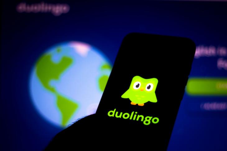 This image portrays the Duolingo app logo on a smartphone with the homepage of the app open on a screen in the background