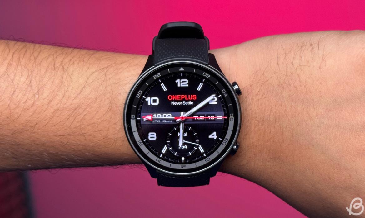 OnePlus 2R on a person's wrist with an analog watch face in front of a bright pink background